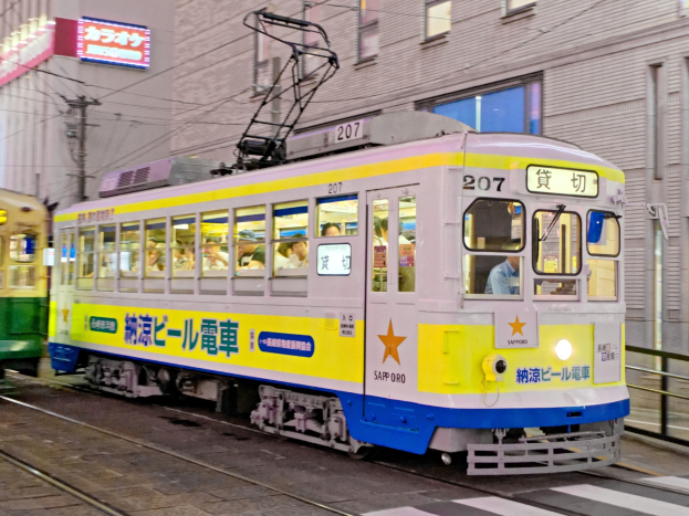 Eine gelbe und blaue Straßenbahn auf einer Stadtstraße in der Nacht mit Menschen darin, Gebäude mit Fenstern, Strommasten mit Drähten und Schilder im Hintergrund.