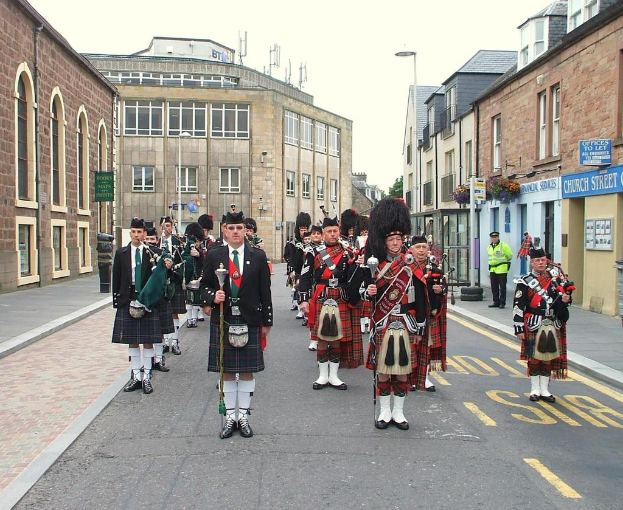 Eine Gruppe von Menschen, einige stehen auf der Straße und andere spielen Musikinstrumente, mit Gebäuden, Plakaten, Pfählen, Bäumen und Reifen im Hintergrund.