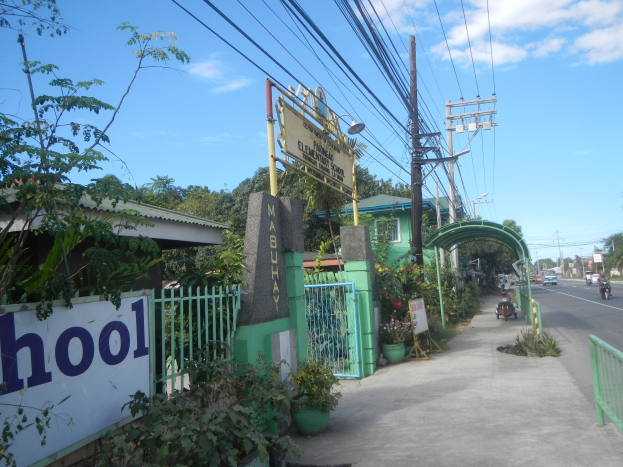 Schulhof mit einem Schild am Straßenrand mit einem Bogen, Tor, Namensschild, Fahrzeugen, Pflanzen, Bäumen, Strommasten mit Drähten und einem bewölkten Himmel im Hintergrund.