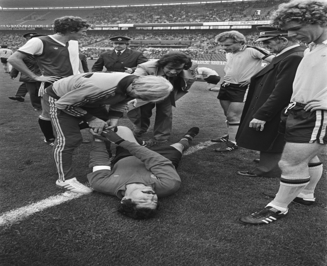 Schwarzes und weißes Bild eines Fußballspielers, der auf dem Boden liegt und von einer Gruppe Männer umgeben ist, mit einer Zuschauermenge im Hintergrund des Stadions.