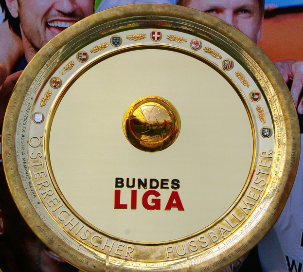 Ein Mann hält einen goldenen Schild mit der Aufschrift 'Bundesliga', im Hintergrund eine Fahne mit drei Personen.