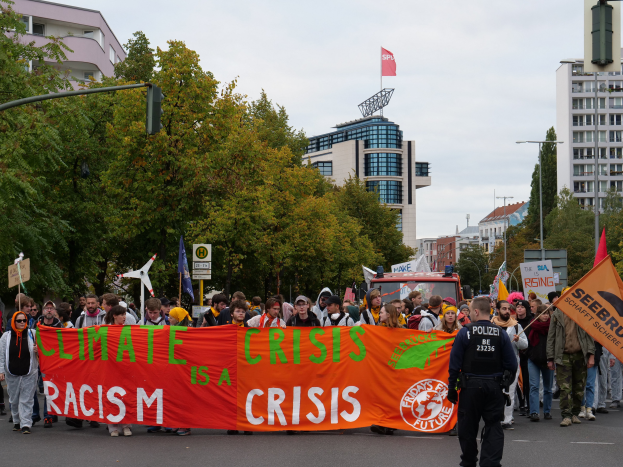Eine Gruppe von Menschen marschiert eine von Bäumen gesäumte Straße entlang und hält ein Schild hoch, auf dem "Klimakrise ist eine Krise" steht, mit Gebäuden und einem klaren blauen Himmel im Hintergrund.