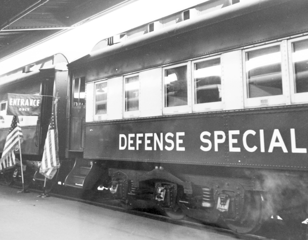 Schwarz-weiß-Foto eines Zuges mit der Aufschrift "Defense Special" auf einem Bahngleis mit zwei Flaggen links und einer Decke oben.
