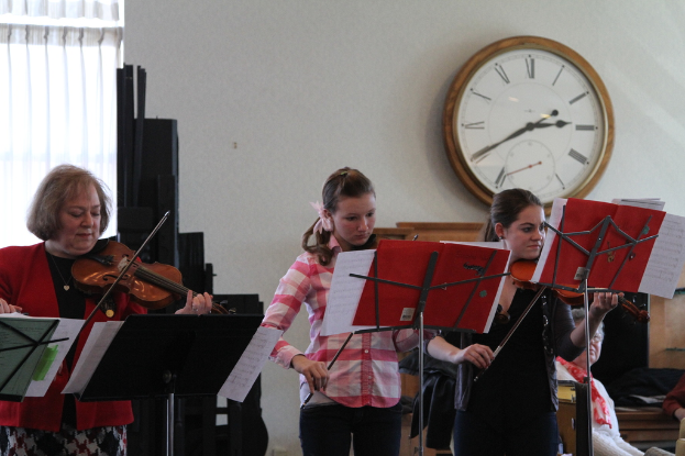 Drei Frauen auf der Bühne mit Violinen, mit Notenständern und Noten davor, während ein Zuschauer im Vordergrund sitzt; eine Uhr ist an der weißen Wand im Hintergrund angebracht und ein weißer Vorhang ist links zu sehen.