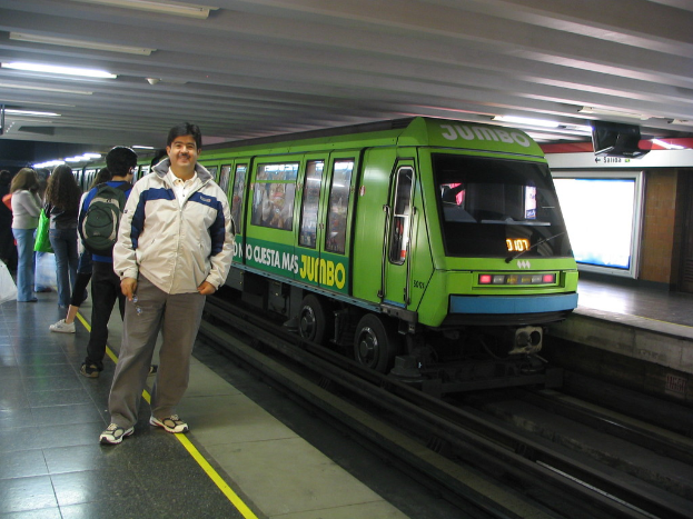 Ein Zug auf einer Eisenbahnschiene mit Menschen, die links stehen und ein Dach oben im Bild.