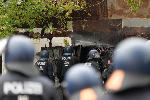Gruppe von Polizisten in schwarzen Uniformen und Helmen, die vor einem Gebäude mit Maschendrahtzaun und Bäumen im Hintergrund stehen.