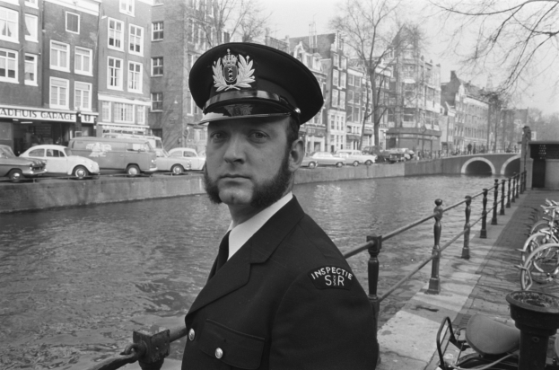 Ein Mann in Polizeiuniform steht neben einem Fluss, mit Fahrrädern auf dem Gehweg zu seiner Rechten, Fahrzeugen auf der Straße, einer Brücke, Bäumen, Gebäuden mit Fenstern und einem klaren Himmel im Hintergrund.