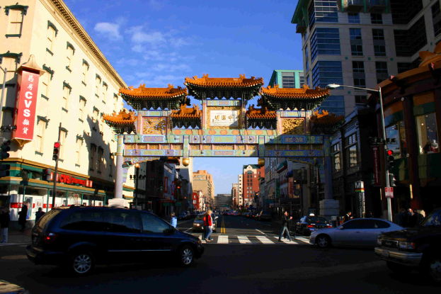 Eine Straße mit fahrenden Fahrzeugen, Fußgängern, die eine Zebrastreifen überqueren, einem Bogen im Hintergrund, Gebäuden auf beiden Seiten und einer Ampel unter einem klaren Himmel.