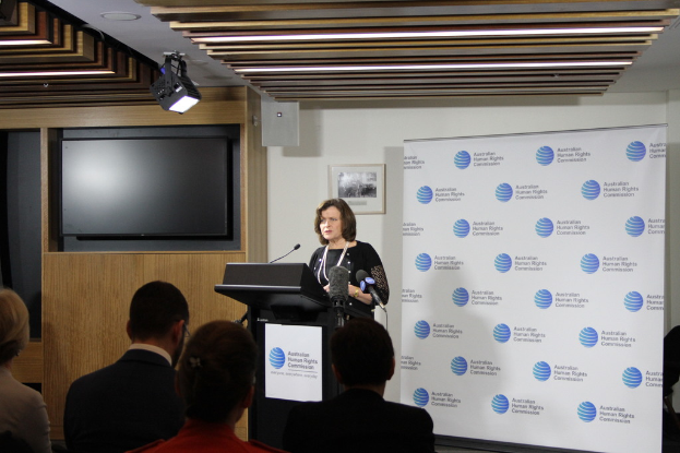 Eine Frau steht auf einer B├╝hne und spricht in ein Mikrofon, mit einem Publikum vor ihr, einem Banner an der Wand hinter ihr, einem Fernseher an der h├Âlzernen Wand und Lichtern an der Decke.