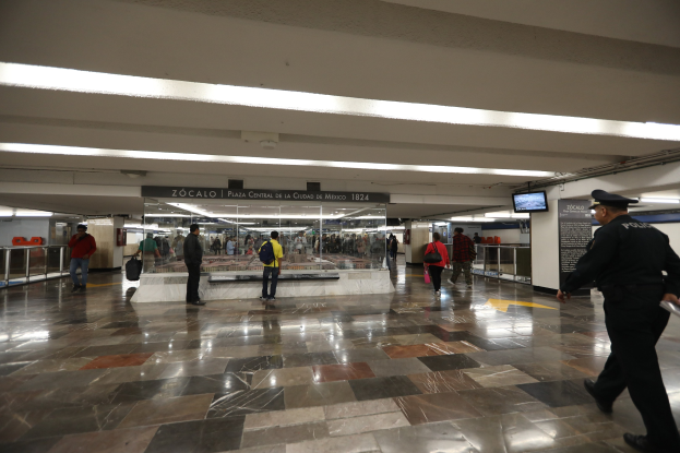 Ein überfüllter U-Bahnhof mit Menschen, die an einem Polizeibeamten im Vordergrund vorbeigehen, mit überkopftexttafeln, Bildschirmen, Lampen, Geländern und verschiedenen Gegenständen.