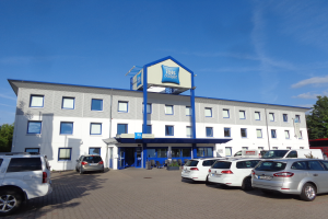 Ein Gebäude mit einer Tafel, auf der "ibis budget frankfurt airport" steht, parkende Autos, Topfpflanzen, ein Straßenpfahl, Bäume und ein bewölkter Himmel.