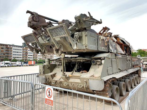 Ein gepanzertes Fahrzeug steht vor einem Metallzaun mit einem daran befestigten Schild, mit Fahrzeugen, Bäumen, Gebäuden und einem klaren blauen Himmel im Hintergrund.