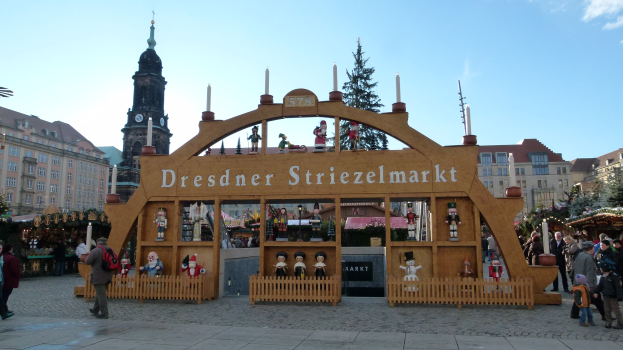 Ein belebter Weihnachtsmarkt in Dresden, Deutschland mit Menschen, die gehen, Taschen tragen und ein Tor im Hintergrund, umgeben von Bäumen, Gebäuden und einem bewölkten Himmel.