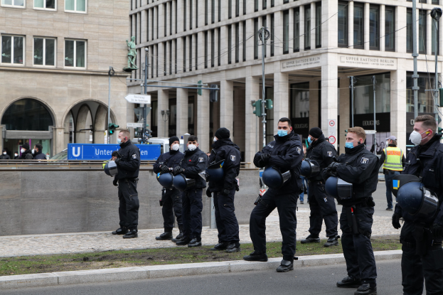 Eine Gruppe von Polizisten in schwarzen Uniformen und Masken steht vor einem Berliner Gebäude mit Glasfenstern und Säulen, einige halten Helme, mit Laternenmasten, Verkehrsampeln, Texttafeln, einer Statue und anderen Menschen im Hintergrund auf einem grasbewachsenen Boden.