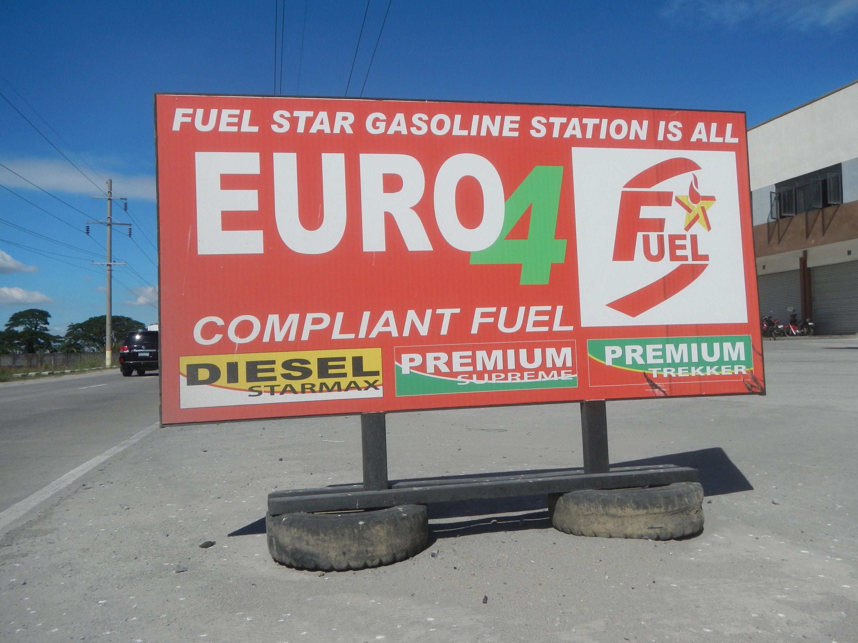 Tankstellen-Schild mit der Aufschrift "Euro4 Fuel" mit Fahrzeugen auf der Straße, Strommästen, Bäumen, einem Gebäude und Himmel im Hintergrund.