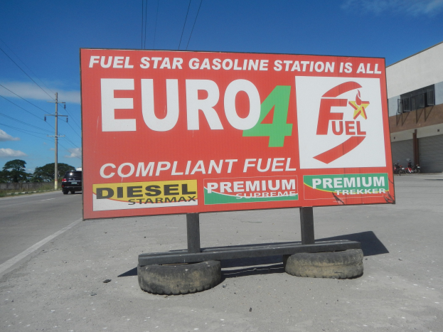 Tankstellen-Schild mit der Aufschrift "Euro4 Fuel" mit Fahrzeugen auf der Straße, Strommästen, Bäumen, einem Gebäude und Himmel im Hintergrund.