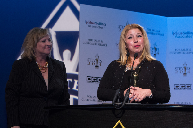 Eine Frau auf der linken Seite, die lächelt, und eine andere Frau in einem schwarzen Kleid, die ein Objekt hält und an einem Podium mit zwei Mikrofonen spricht, mit Bannern im Hintergrund, die Bilder und Text anzeigen.