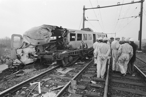 Ein Schwarz-Weiß-Bild einer Zugkollision mit Menschen in Helmen um das Wrack herum, Strommasten, Bäume, Gebäude und einen klaren Himmel im Hintergrund.