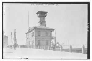 Schwarz-weiß-Foto einer Feuerwache an der Battery, New York, mit einem Gebäude, Menschen, Pfählen, einem Zaun, Booten auf dem Wasser und einem bewölkten Himmel.