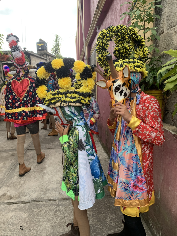 Eine Gruppe von Menschen in bunten Kostümen, einige halten Gegenstände, stehen zusammen vor einem Gebäude während eines Faschingsumzugs, mit Bäumen, Gebäuden und einem klaren blauen Himmel im Hintergrund.