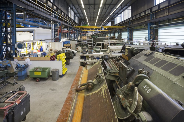 Ein großes Lagerhaus voller Maschinen, einschließlich eines Landfahrzeugs auf der rechten Seite, Metallobjekten auf der linken Seite und Lampen oben.