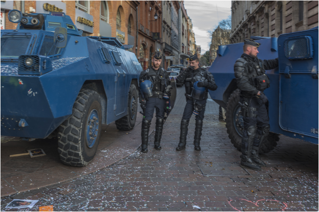 Eine Gruppe von Polizisten steht vor einem gepanzerten Fahrzeug auf einer Straße, mit Gebäuden, Bäumen und einem bewölkten Himmel im Hintergrund.