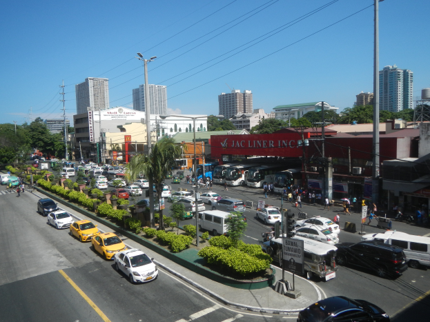 Eine belebte Stadtstraße mit zahlreichen Autos, Bussen und Fußgängern, umgeben von Gebäuden, Bäumen und diverser städtischer Infrastruktur unter einem klaren blauen Himmel.