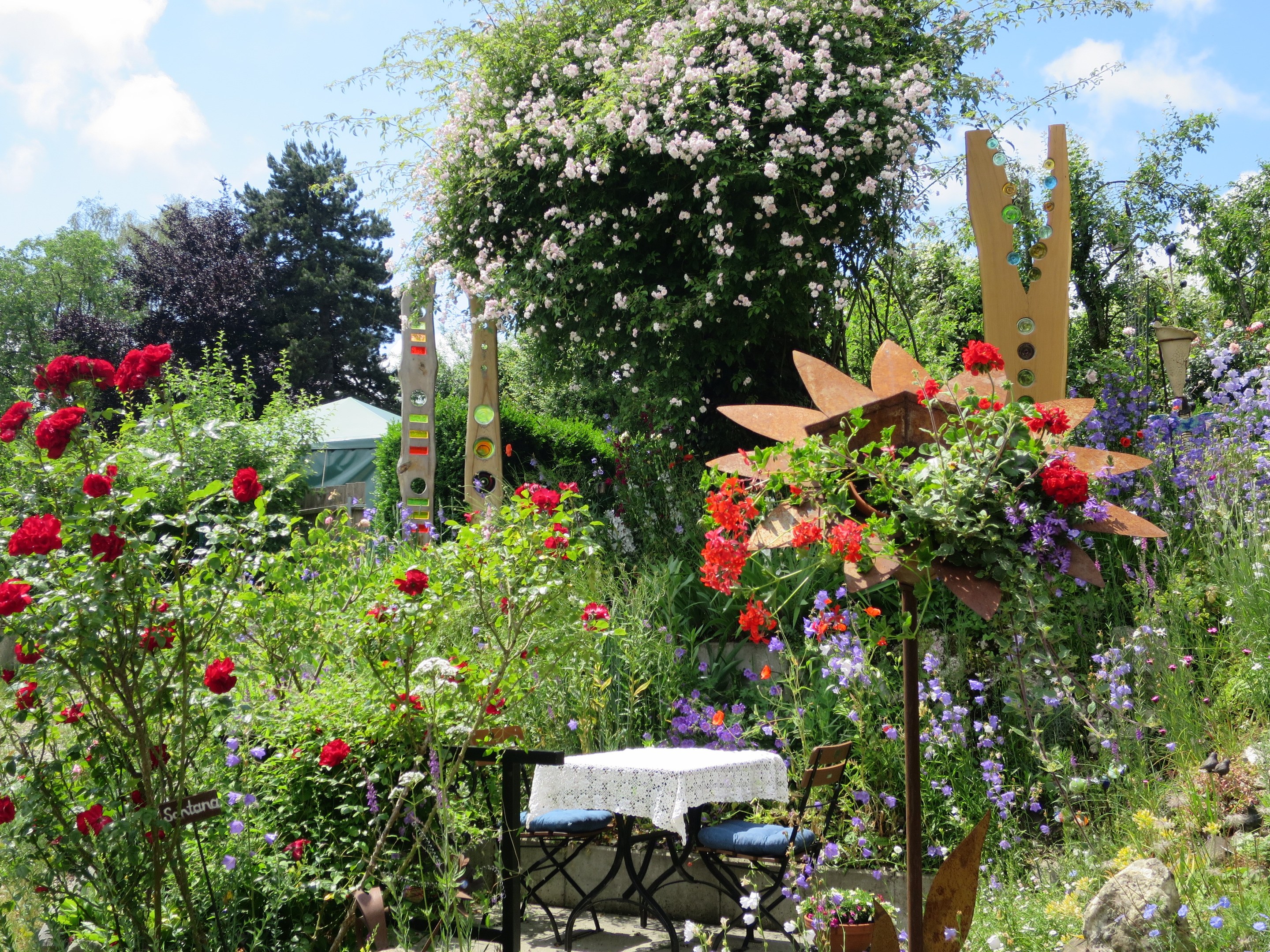 Ein Garten mit bunten Blumen und Pflanzen, der einen Tisch mit einer Decke und Stühlen zeigt, umgeben von Bäumen unter einem bewölkten Himmel auf der Chelsea Flower Show.