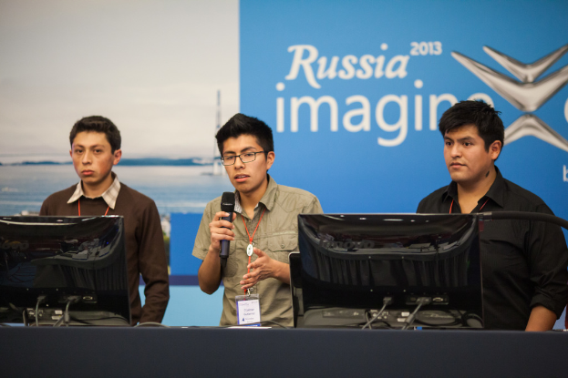 Drei Jungen stehen neben einem Schreibtisch mit zwei Computersystemen, einer spricht in ein Mikrofon, trägt ID-Karten mit roten Markierungen, vor einer Wand mit einem Werbeplakat, auf dem "Russia Imagine 2013" steht.