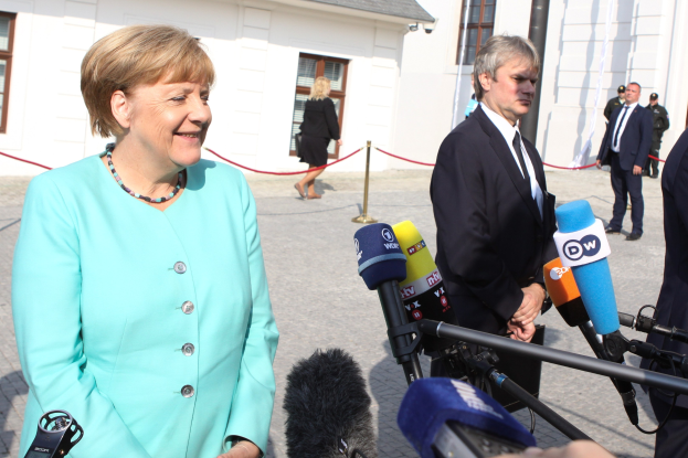 Bundeskanzlerin Angela Merkel beim Sprechen mit Reportern vor einem Gebäude mit einer Gruppe von Menschen und Equipment unter einem bewölkten Himmel.