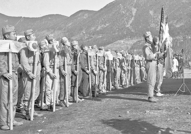Ein Schwarz-Weiß-Bild von Menschen in Uniformen, die Fahnen halten und auf einem steinigen Pfad mit Hügeln im Hintergrund und einem sichtbaren Himmel stehen.