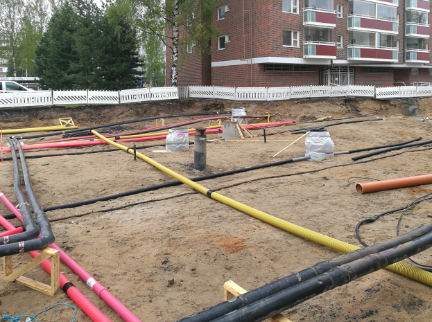 Großer Haufen von Rohren auf einer Baustelle mit Sand, hölzernen Gegenständen, Folienabdeckungen und anderen Materialien im Vordergrund, Bäume und Gebäude im Hintergrund unter einem klaren blauen Himmel.