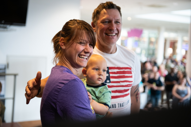 Ein Mann in einem weißen T-Shirt und eine Frau mit einem Baby stehen vor einer Menge mit einem Fernseher an der Wand im Hintergrund.