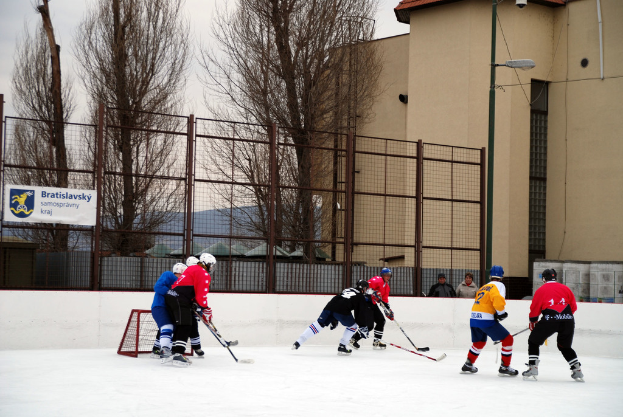 Personen beim Eishockey auf einem Eisring mit Gebäuden, Bäumen, einer Straßenlaterne, einem Schild und Zäunen im Hintergrund unter einem Himmel.