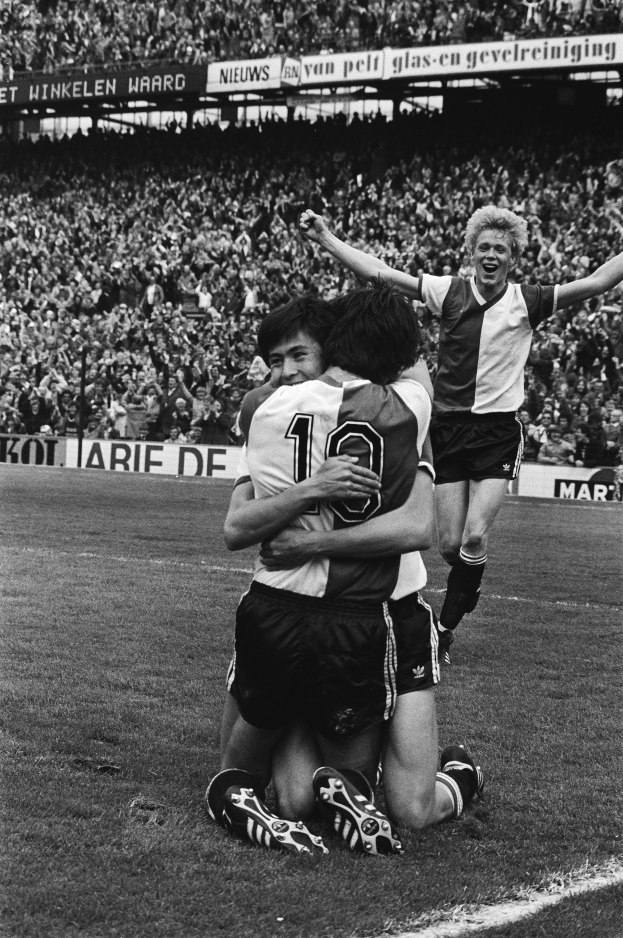 Drei Fussballspieler feiern ein Tor auf dem Boden vor einer jubelnden Menge in einer schwarz-weissen Stadion-Szene.