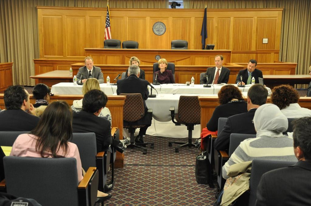 Ein Konferenzraum mit einer Gruppe von Menschen, die auf Stühlen sitzen, zwei ausländische Flaggen, ein orangefarbener Schreibtisch mit Wasserflaschen und einem Mikrofon.