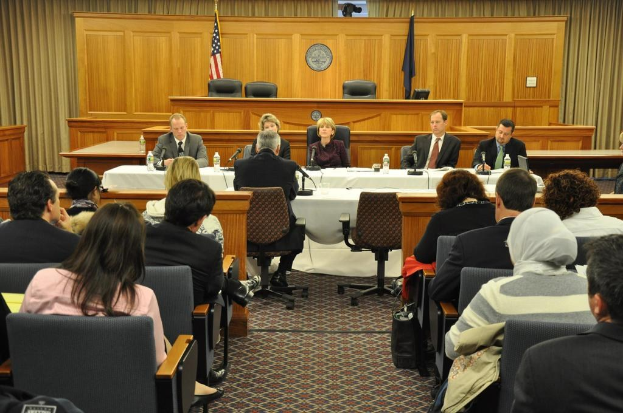 Ein Konferenzraum mit einer Gruppe von Menschen, die auf Stühlen sitzen, zwei ausländische Flaggen, ein orangefarbener Schreibtisch mit Wasserflaschen und einem Mikrofon.