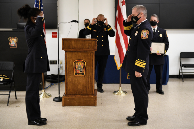 Gruppe uniformierter Personen auf einem Podium während einer Vereidigung eines neuen Feuerwehrchefs, der eine Urkunde hält, mit Flaggen und Sitzplätzen im Hintergrund.
