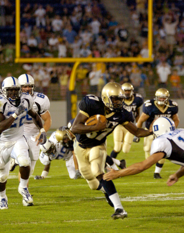 Ein Fussballspieler in Sportkleidung und Helm rennt mit dem Ball, umgeben von anderen Spielern, mit unscharfem Hintergrund und Text unten.