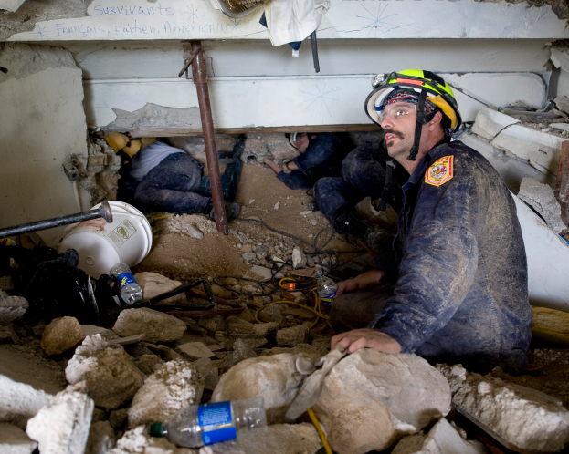 Ein Feuerwehrmann in einem Helm sitzt zwischen Trümmern, darunter Steine und Flaschen, mit einer teilweise sichtbaren Wand im Hintergrund, die wahrscheinlich den Namen des Gebäudes anzeigt, das von einem israelischen Luftangriff getroffen wurde.