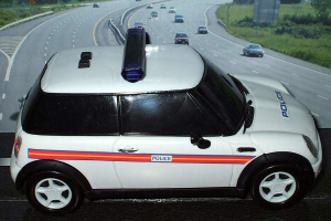 Weißes Polizeiauto auf einer Straße mit mehreren Fahrzeugen, Gras und Bäumen auf beiden Seiten, fotografiert ausserhalb einer Stadt.