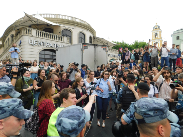 Eine große Gruppe von Menschen steht vor einem Gebäude, einige halten Kameras und Handys, mit einem geparkten Fahrzeug, Bäumen und einem klaren blauen Himmel im Hintergrund, was auf eine Protestaktion gegen das russische Drohnenverbot hindeutet.