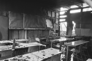 Schwarzes und weißes Foto eines verlassenen Klassenzimmers mit verkohlten Wänden, zerbrochenen Fenstern und verstreuten Tischen und Stühlen.