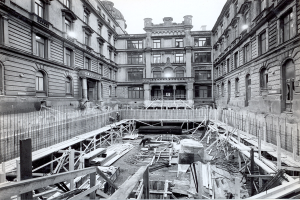 Schwarzes und weißes Foto einer Baustelle in einer Stadt mit Gebäuden im Hintergrund, hölzernen Gegenständen und Metallstangen im Vordergrund und Text unten rechts.