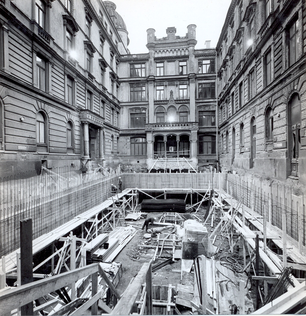 Schwarzes und weißes Foto einer Baustelle in einer Stadt mit Gebäuden im Hintergrund, hölzernen Gegenständen und Metallstangen im Vordergrund und Text unten rechts.
