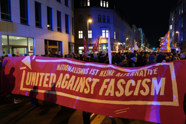 Gruppe von Menschen marschiert mit einem Banner 'Einheit gegen Faschismus' die Straße entlang, mit Gebäuden, Laternenmasten und Schildern im Hintergrund.