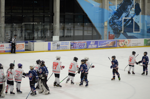 Gruppe junger Menschen beim Eisschnelllaufen auf einer Indoor-Eisbahn, die Helme tragen und Stöcke halten, mit einer Person links daneben und Bannern, Säulen und Glaswänden im Hintergrund.