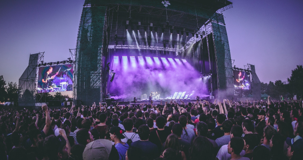 Eine gro├če Menge bei einem Musikfestival schaut auf einer hell erleuchteten Bühne mit Instrumenten und Bildschirmen, vor einer Kulisse aus B├Ąumen, einem Geb├Ąude und einem klaren blauen Himmel.