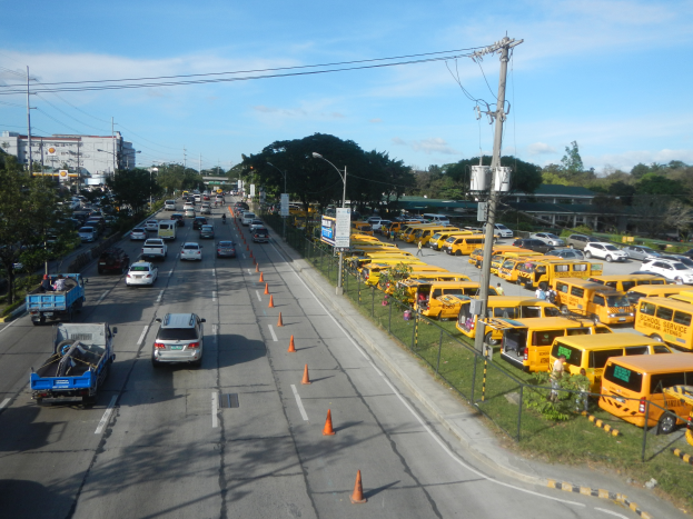 Eine belebte Straßenszene mit verschiedenen Fahrzeugen, Verkehrskegeln, Laternenmasten, Strommasten mit Drähten und Texttafeln, mit einem Maschendrahtzaun auf der rechten Seite, Bäumen, Gebäuden und einem bewölkten Himmel im Hintergrund.