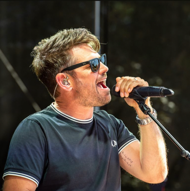Ein Mann in einem schwarzen T-Shirt und Sonnenbrille singt in ein Mikrofon mit Ständer vor einem unscharfen, träumerischen Hintergrund.